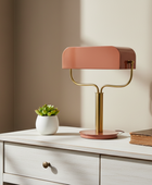 Pink table lamp on a white dresser with a plant and book in a softly lit room.
