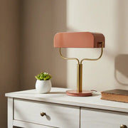 Modern table lamp with a pink shade and gold base on a white surface with a plant and book.
