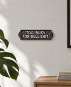 A rectangular cast iron sign with the text 'TOO BUSY FOR BULL SHIT' in raised white lettering, mounted on a dark gray surface.
