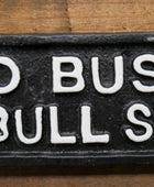 A close-up of a black rectangular sign or plaque with raised white lettering that reads "O BUST BULL SH" on a wooden surface.