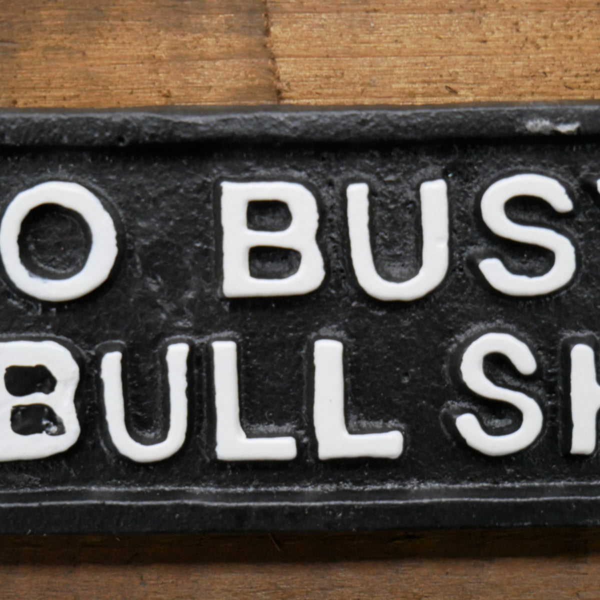 A close-up of a black rectangular sign or plaque with raised white lettering that reads "O BUST BULL SH" on a wooden surface.