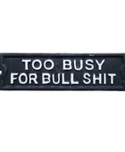 A rectangular black cast iron sign with raised white lettering that reads 'TOO BUSY FOR BULL SHIT', photographed against a rustic wooden surface.