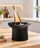 A black top-hat-shaped ice bucket on a wooden kitchen counter, holding a sparkling wine bottle and ice. Two filled champagne flutes, a bowl of green grapes, and a cutting board with greenery sit to the left, with a bright modern kitchen in the background.