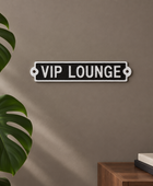 A rectangular black wall sign with a white border and the words 'VIP LOUNGE' in white uppercase letters, mounted on a taupe wall. A large green monstera leaf appears on the left, and a wooden side table with stacked books and a dark bowl is visible on the right.