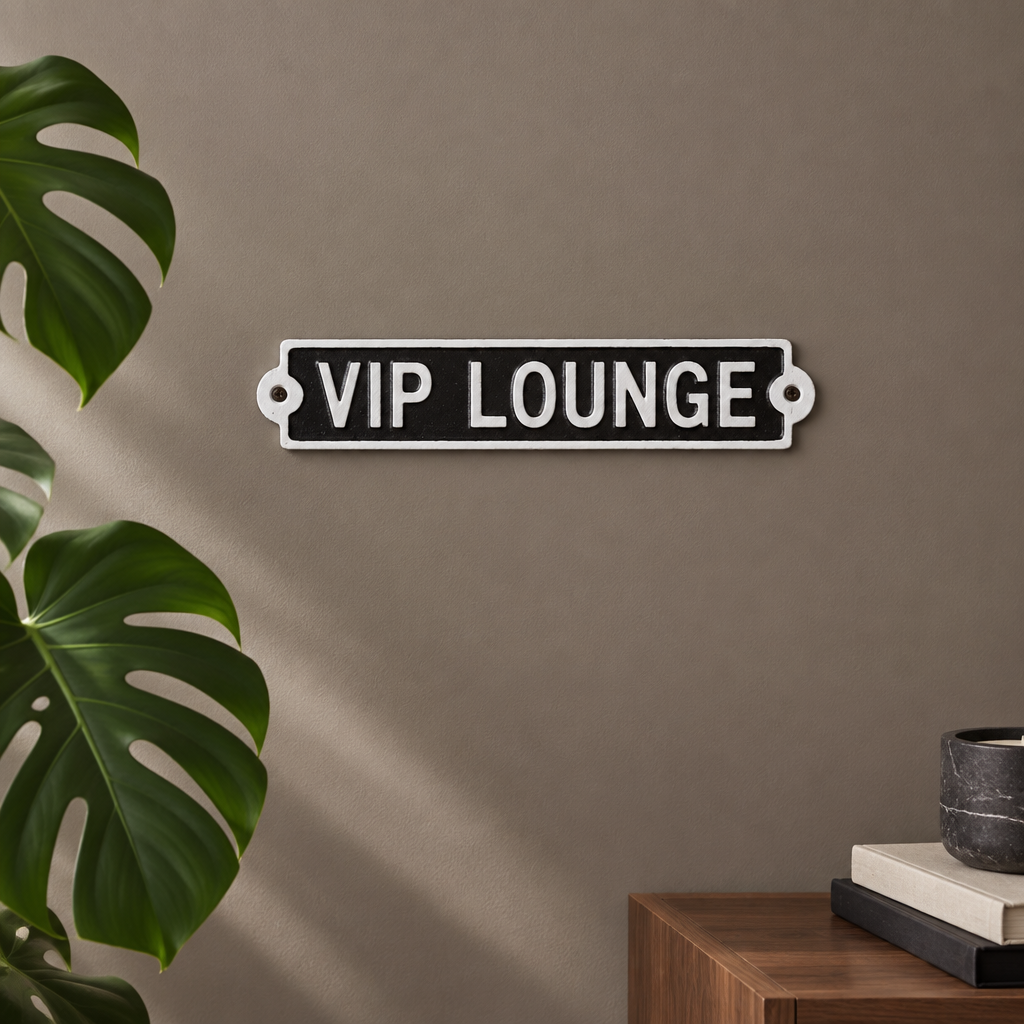 A rectangular black wall sign with a white border and the words 'VIP LOUNGE' in white uppercase letters, mounted on a taupe wall. A large green monstera leaf appears on the left, and a wooden side table with stacked books and a dark bowl is visible on the right.
