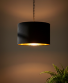 A black drum pendant light with a warm gold interior glow, hanging from a black cord against a dark brown background with a green fern in the lower right corner.