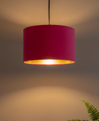 A modern drum pendant light with a vibrant magenta pink fabric shade and a polished gold interior, suspended against a warm gray wall with a soft glow.