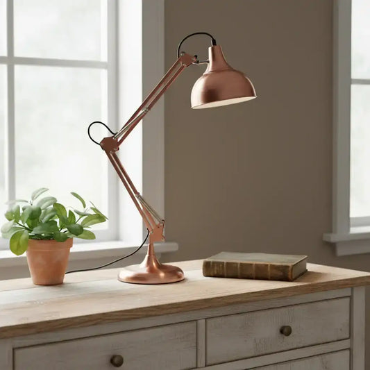 Copper desk lamp on a wooden surface with a plant and book in a room with windows.