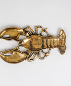 A metallic lobster-shaped decorative object with a warm gold/brass finish, photographed on a plain light gray background.
