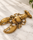 A lobster-shaped decorative object in a metallic antique gold finish, resting on a white and gray marble surface with soft diagonal shadows.