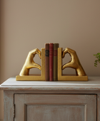 A pair of gold bookends shaped like hands forming a heart with their fingers, holding a set of three books on a light wood console table next to a potted green plant.