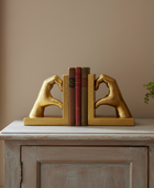 A pair of gold decorative bookends shaped like hands forming heart gestures, holding a set of books on a distressed wood cabinet against a beige wall.