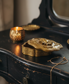 An ornate gold-toned jewelry box with a carved lid sits on a distressed black wooden vanity table next to a lit candle holder and a gold chain necklace.