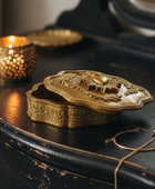 An ornate brass jewelry box with a floral lid sits on a distressed black wooden surface next to a lit candle holder and a delicate gold chain.
