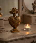 An ornate antique-style brass heart-shaped urn with a flame-like lid, displayed on a distressed gray wooden dresser next to a lit candle in a glass holder.