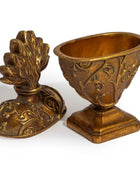 An ornate antique-style bronze decorative object shown in two pieces, with one half standing as a pedestal base and the other half leaning against it to reveal a sculpted floral or leafy top detail, both decorated with embossed scrollwork.