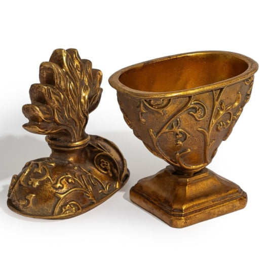An ornate antique-style bronze decorative object shown in two pieces, with one half standing as a pedestal base and the other half leaning against it to reveal a sculpted floral or leafy top detail, both decorated with embossed scrollwork.