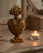 An ornate antique-style bronze heart-shaped urn with a floral lid, displayed on a rustic wooden surface next to a lit candle and a gold bracelet.