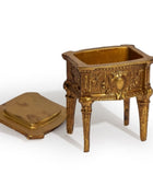 An ornate miniature gold-toned metal candelabra or decorative pedestal with a matching lid placed beside it, photographed on a clean white background.