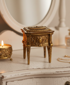 An ornate gold-toned jewelry box with carved detailing stands on a distressed white vanity table next to a lit candle, a small perfume bottle, and a gold chain bracelet.