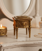 An ornate antique-style gold-toned jewelry box with carved floral details and four legs, displayed on a distressed white dresser next to a lit candle and a perfume bottle.