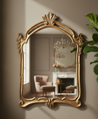 An ornate gold-framed wall mirror with classical baroque-style scrollwork and a leaf crest at the top, mounted on a beige wall. The mirror reflects a softly lit interior with a chair and fireplace, and a green plant is visible on the right side.