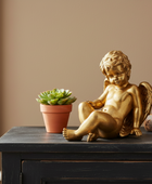 Gold cherub figurine and potted succulent on a dark surface with a beige wall background