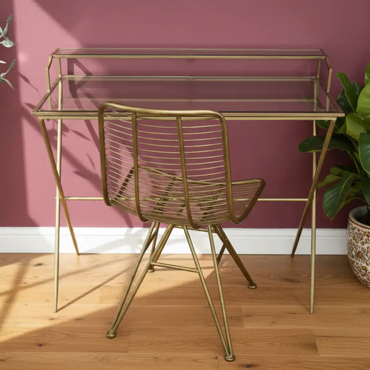 Gold metal desk with glass top and matching chair against a light gray wall.
