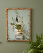 Framed artwork of birds and foliage on a green wall with a plant in the foreground.