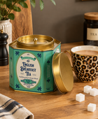A teal and gold metal tin of English Breakfast Tea sits on a wooden table next to a leopard-print mug filled with tea, with sugar cubes, a plant, and kitchen props in the background.