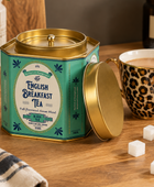 A teal and gold hexagonal metal tea tin labeled English Breakfast Tea, shown on a wooden table beside a leopard-print mug of tea, sugar cubes, and a pepper mill.