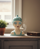 Decorative head sculpture with a blue hat on a wooden surface next to a plant and book.