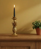 Gold pineapple candle holder with a lit candle on a wooden surface against a beige wall.