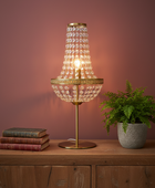 A table lamp with a gold brass stand and a clear crystal prisme shade, glowing with a warm light. It is styled on a wooden cabinet next to a potted fern and a stack of books against a dusty rose wall.