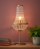 A gold table lamp with a tiered crystal chandelier shade, lit with a warm glow, set on a wooden table next to a potted fern and a stack of vintage books against a dusty rose wall.