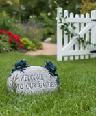 Decorative stone with frogs and 'Welcome to Our Garden' text in a garden setting with flowers and a white picket fence.