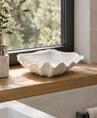 A white ceramic shell-shaped vessel with a textured, ribbed exterior sits on a wooden window sill in a modern bathroom setting.