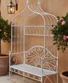 Decorative white metal bench with intricate designs against a beige wall with potted plants.
