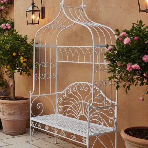 Decorative white metal bench with intricate designs against a beige wall with potted plants.