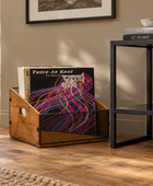 A wooden record bin holding vinyl records on a hardwood floor in a styled room, with a potted plant, framed wall art, and a black metal side table in the background.