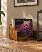 A wooden and black mesh record rack holding a vinyl record, placed on a wood floor in a styled room with a plant, framed wall art, and a side table visible.