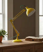 Yellow adjustable desk lamp on a wooden surface with a plant and book in the background