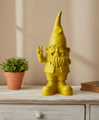 Yellow gnome figurine making a peace sign on a wooden surface with a plant and book in the background.