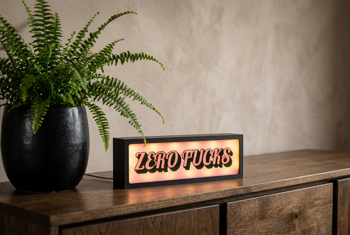 A rectangular black LED sign with the text 'ZERO FUCKS' glowing in warm orange and pink tones, placed on a wooden cabinet next to a potted green fern against a textured beige wall.