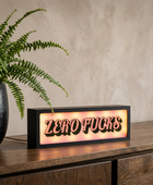 A rectangular black LED sign with the words 'ZERO FUCKS' in raised red letters, glowing with a warm light, placed on a wooden cabinet next to a potted fern against a textured beige wall.