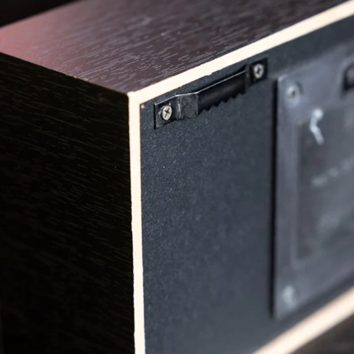 Close-up of the side and back corner of a rectangular wooden box or speaker-like device, showing a dark wood grain exterior, a black textured panel, screws, and a mounting area.