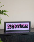 A rectangular novelty sign with the words 'ZERO FUCKS' in bold pink letters outlined in black, set against a light lavender background and mounted on a dark wooden surface.