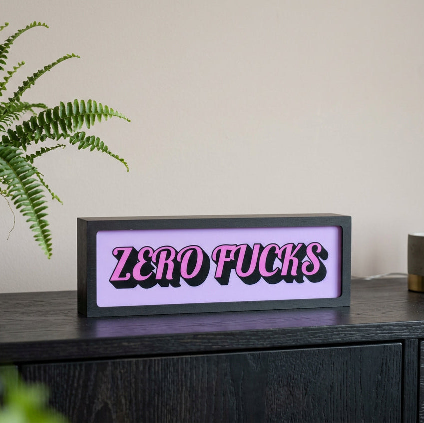 A rectangular novelty sign with the words 'ZERO FUCKS' in bold pink letters outlined in black, set against a light lavender background and mounted on a dark wooden surface.
