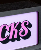 A close-up of a black-framed sign or plaque with a glowing light panel. The panel shows the word "BUCKS" in stylized pink letters with black drop shadows on a pale pink-lilac background.
