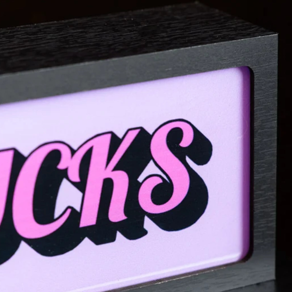 A close-up of a black-framed sign or plaque with a glowing light panel. The panel shows the word "BUCKS" in stylized pink letters with black drop shadows on a pale pink-lilac background.